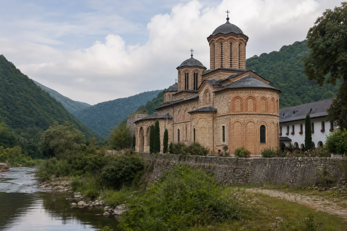 Mănăstire ortodoxă medievală din piatră și cărămidă pe malul unui râu, la poalele munților împăduriți