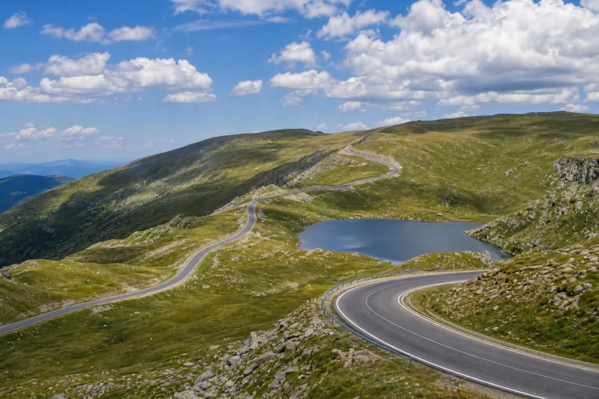 Porțiune alpină de mare altitudine a Transalpinei în nordul Vâlcii, cu culmi verzi și un lac mic lângă drum